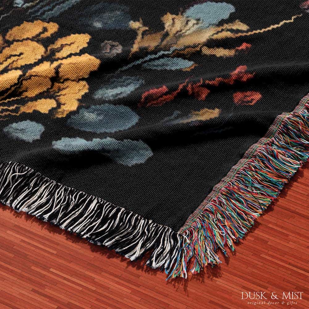 Woven cotton blanket featuring a botanical-style cluster of mushrooms in red, gold, and purple caps surrounded by plants and foliage on a black background.