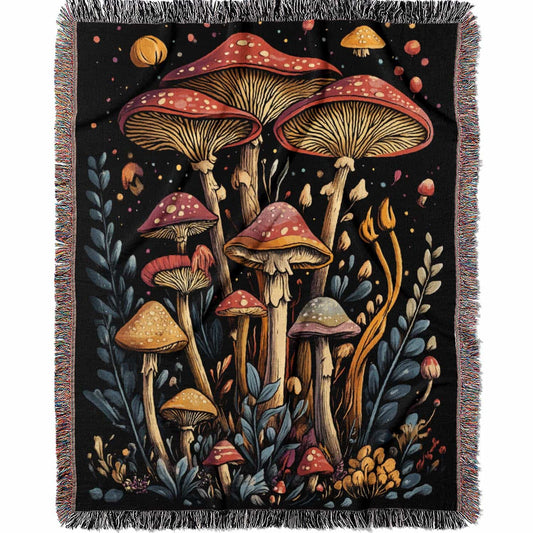 Woven cotton blanket featuring a botanical-style cluster of mushrooms in red, gold, and purple caps surrounded by plants and foliage on a black background.