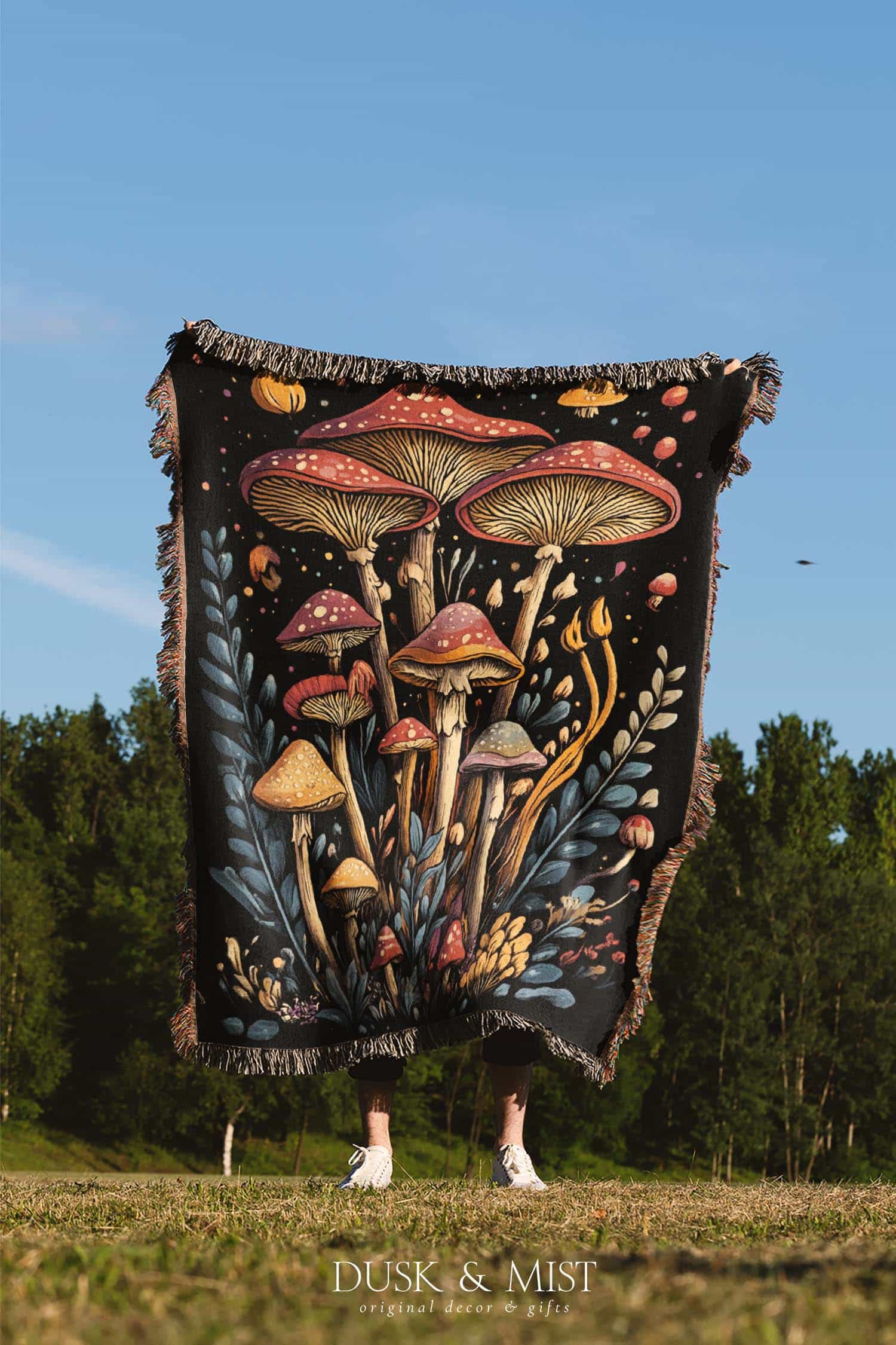 Woven cotton blanket featuring a botanical-style cluster of mushrooms in red, gold, and purple caps surrounded by plants and foliage on a black background.