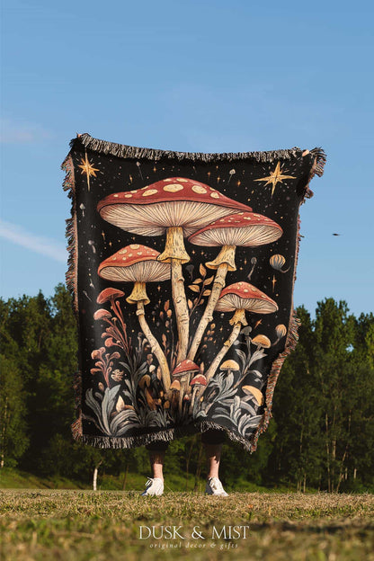 Woven cotton blanket featuring tall red-capped mushrooms under a starry sky with golden stars and surrounding plants on a black background.
