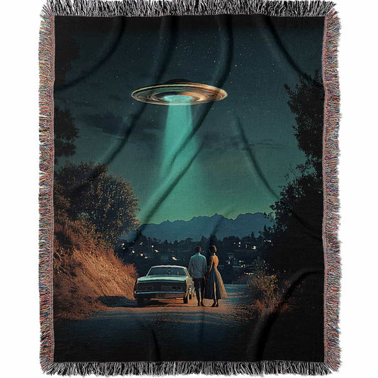 Woven cotton blanket featuring a UFO beaming light onto a couple standing near a car at night, with a starry sky and silhouetted trees.
