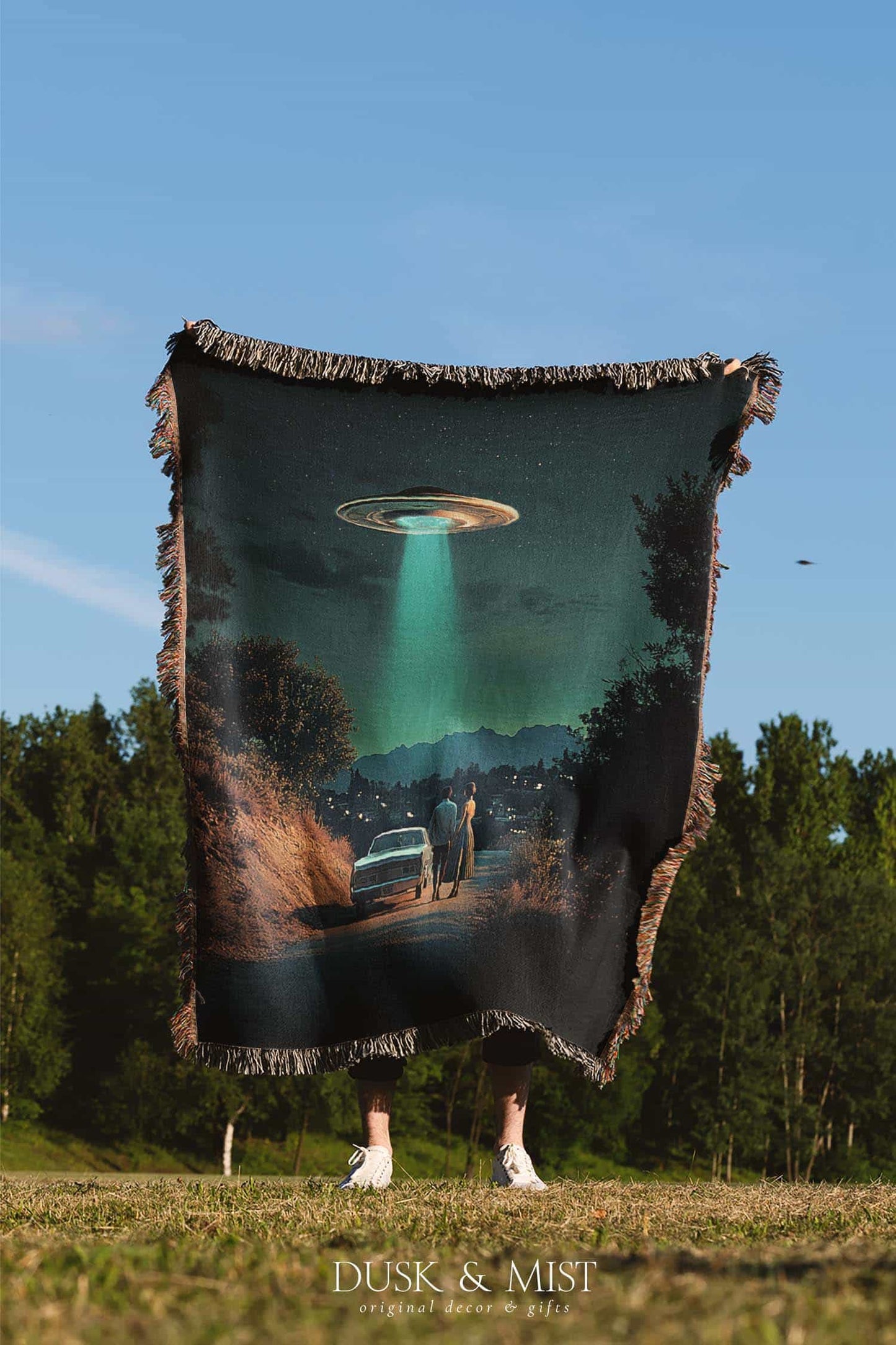 Woven cotton blanket featuring a UFO beaming light onto a couple standing near a car at night, with a starry sky and silhouetted trees.