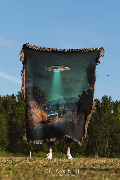 Woven cotton blanket featuring a UFO beaming light onto a couple standing near a car at night, with a starry sky and silhouetted trees.