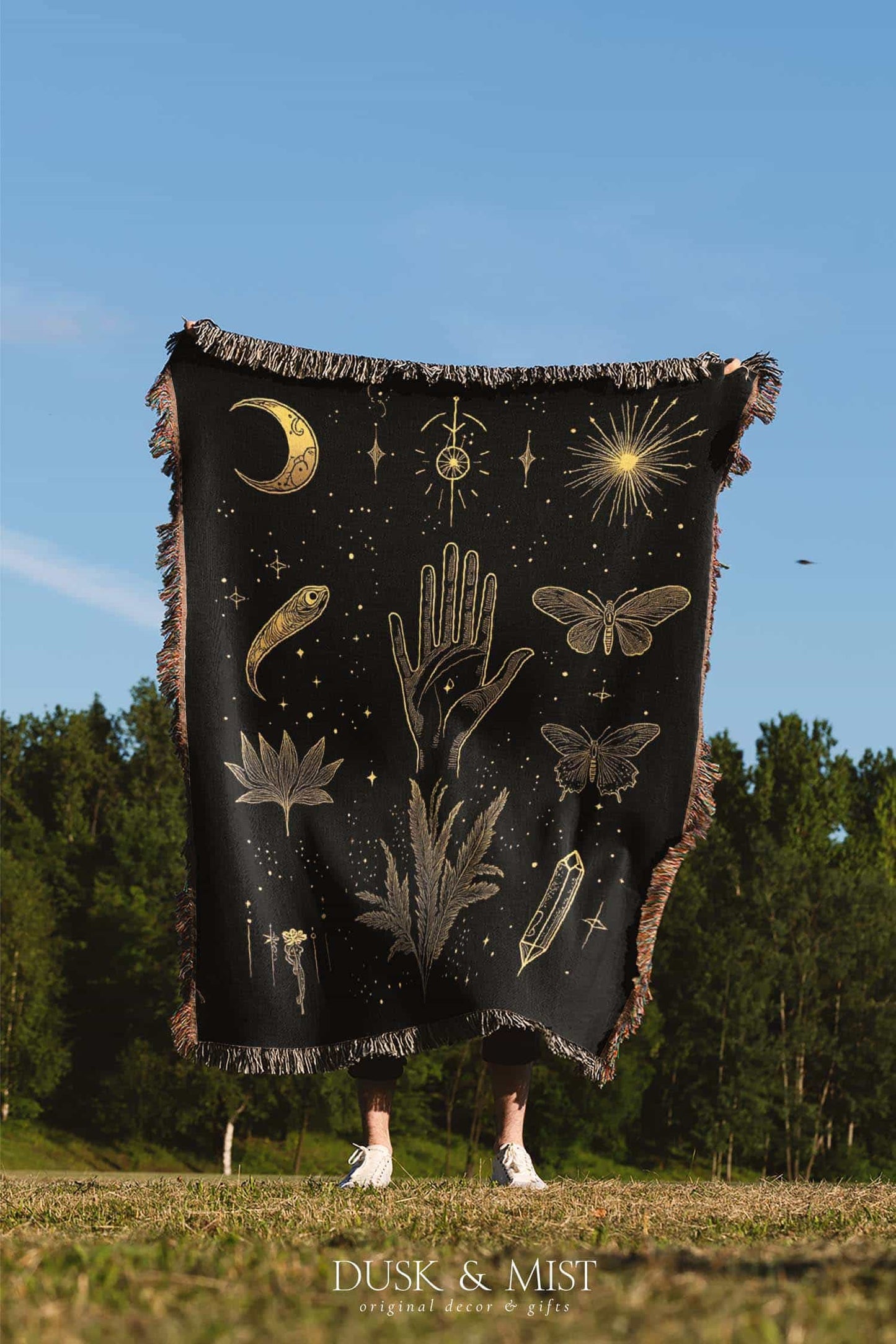 Woven cotton blanket with golden occult illustrations — palmistry hand, crescent moon, moth, snake, crystals, plants, and celestial symbols on a black background.