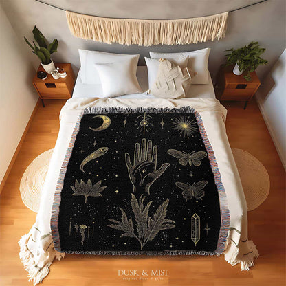 Woven cotton blanket with golden occult illustrations — palmistry hand, crescent moon, moth, snake, crystals, plants, and celestial symbols on a black background.