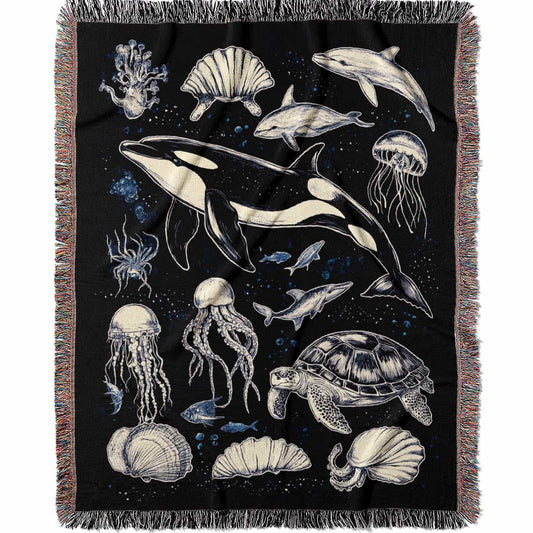 Woven cotton blanket featuring detailed marine life illustration — orca, dolphins, sea turtle, octopus, jellyfish, seashells, and fish on a black background.
