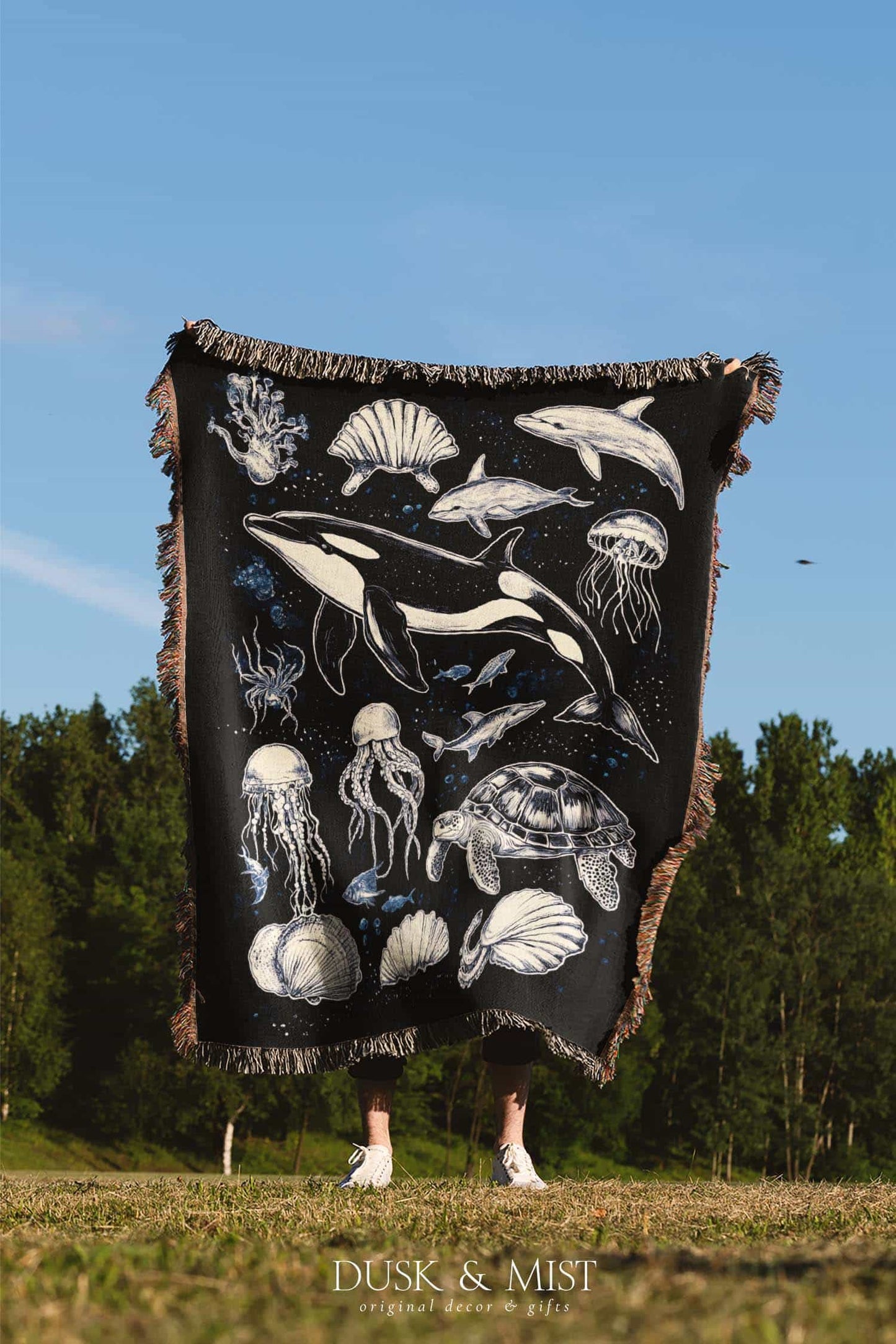 Woven cotton blanket featuring detailed marine life illustration — orca, dolphins, sea turtle, octopus, jellyfish, seashells, and fish on a black background.