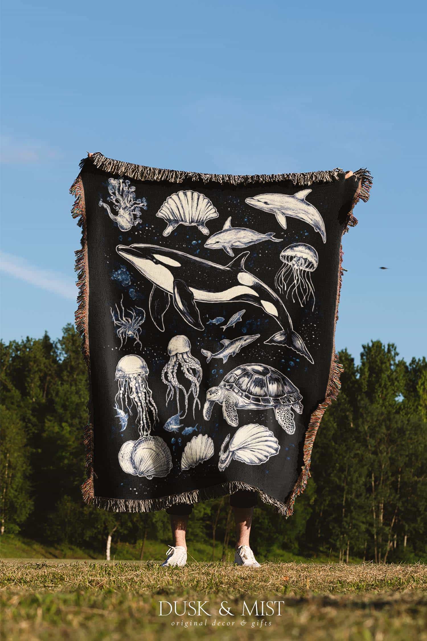 Woven cotton blanket featuring detailed marine life illustration — orca, dolphins, sea turtle, octopus, jellyfish, seashells, and fish on a black background.