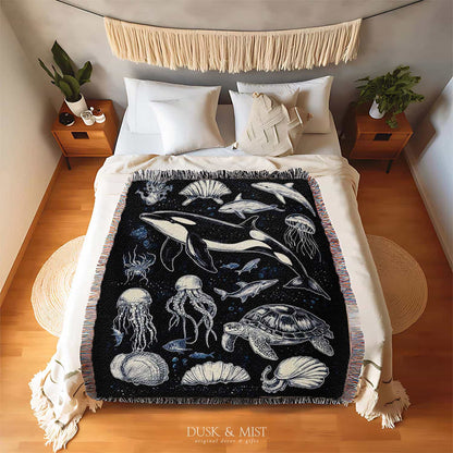 Woven cotton blanket featuring detailed marine life illustration — orca, dolphins, sea turtle, octopus, jellyfish, seashells, and fish on a black background.