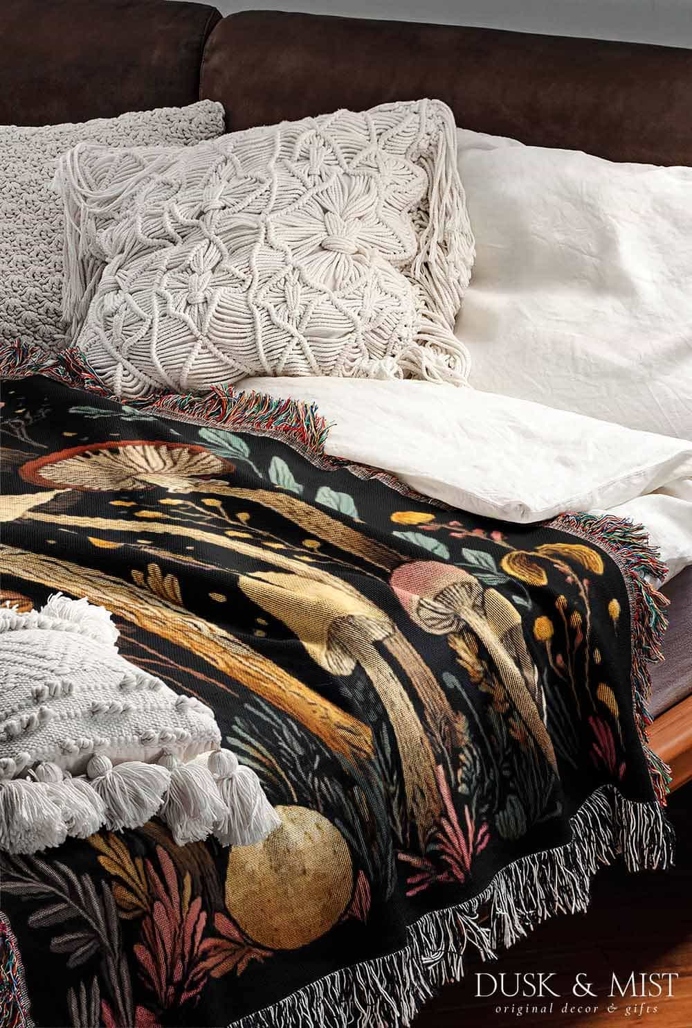 Woven cotton blanket featuring a botanical-style field of mushrooms in red, gold, and cream tones with surrounding plants and foliage on a black background.