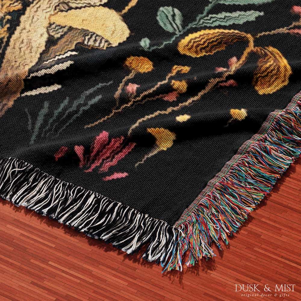 Woven cotton blanket featuring a botanical-style field of mushrooms in red, gold, and cream tones with surrounding plants and foliage on a black background.