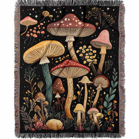 Woven cotton blanket featuring a botanical-style field of mushrooms in red, gold, and cream tones with surrounding plants and foliage on a black background.