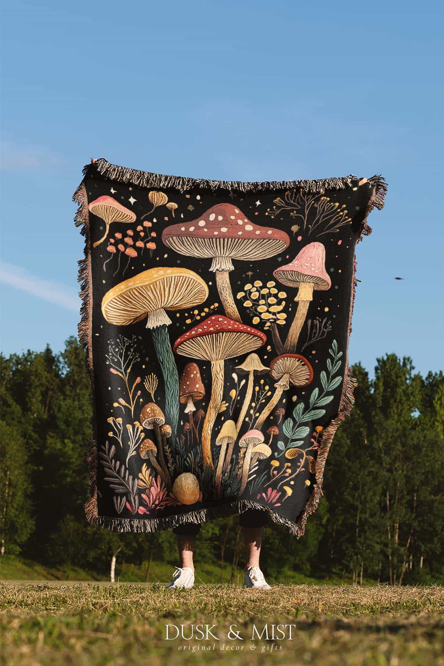 Woven cotton blanket featuring a botanical-style field of mushrooms in red, gold, and cream tones with surrounding plants and foliage on a black background.