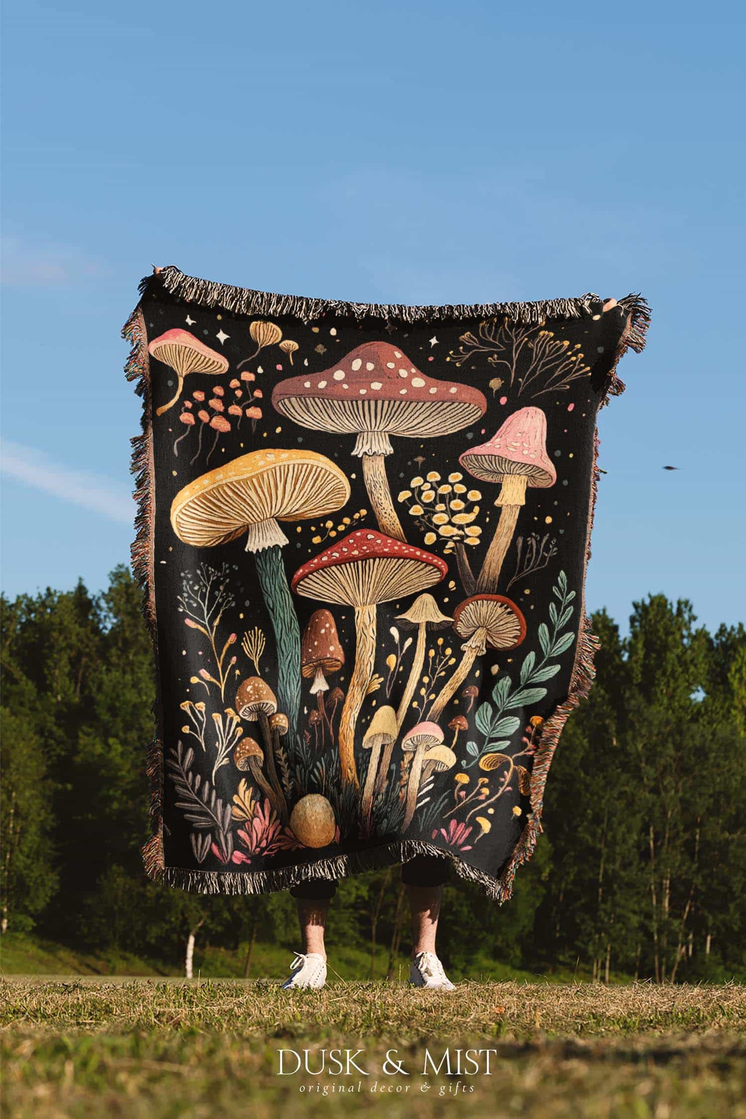Woven cotton blanket featuring a botanical-style field of mushrooms in red, gold, and cream tones with surrounding plants and foliage on a black background.