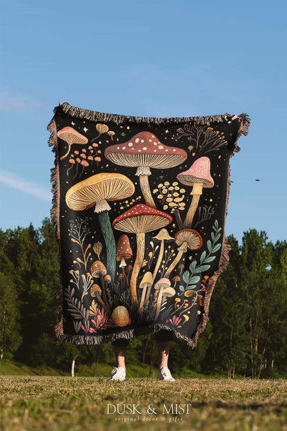 Woven cotton blanket featuring a botanical-style field of mushrooms in red, gold, and cream tones with surrounding plants and foliage on a black background.