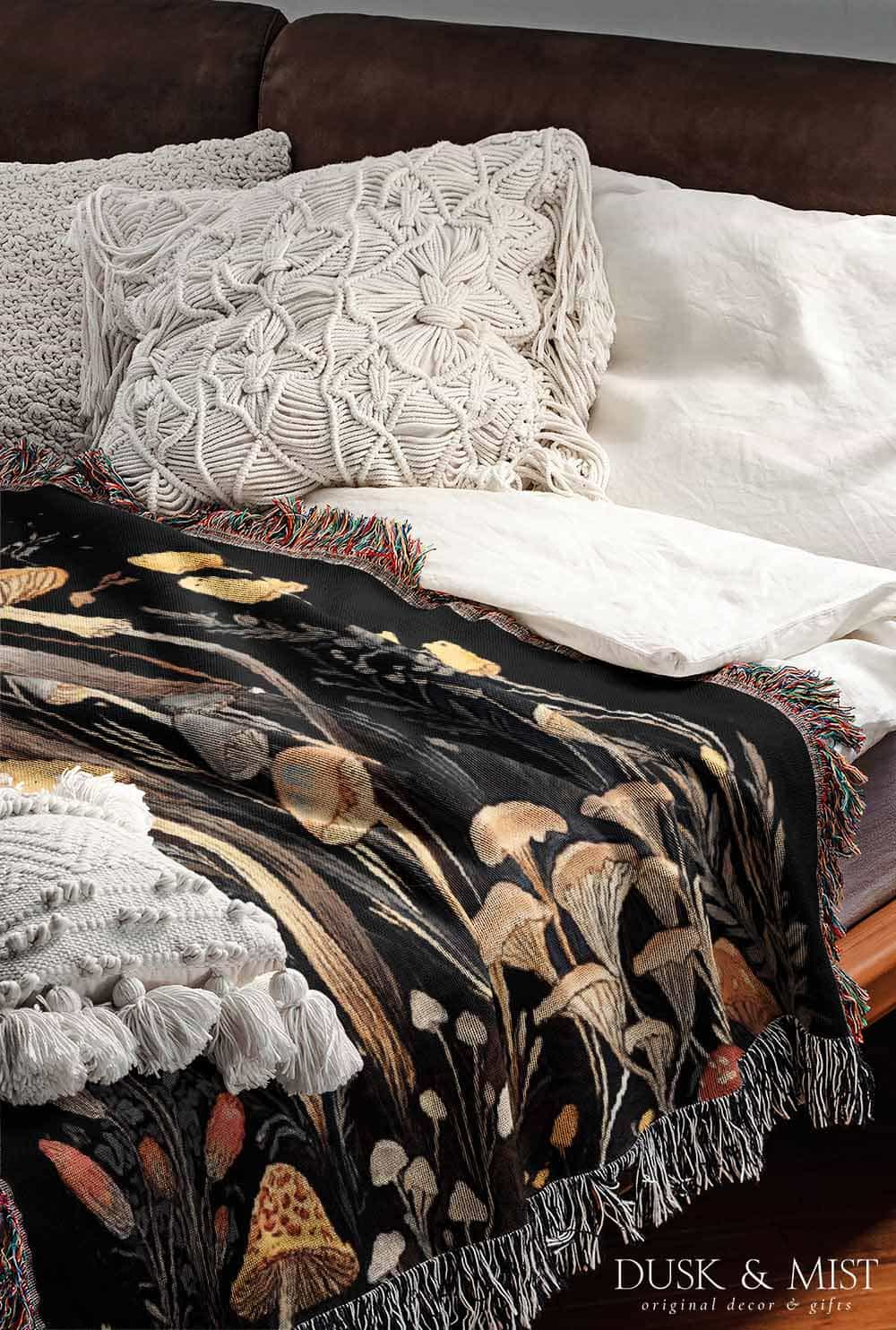 Woven cotton blanket showing a colorful cluster of mushrooms in red, gold, blue, and cream caps with surrounding plants and flowers on a black background.