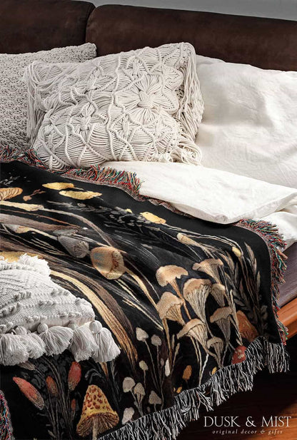 Woven cotton blanket showing a colorful cluster of mushrooms in red, gold, blue, and cream caps with surrounding plants and flowers on a black background.