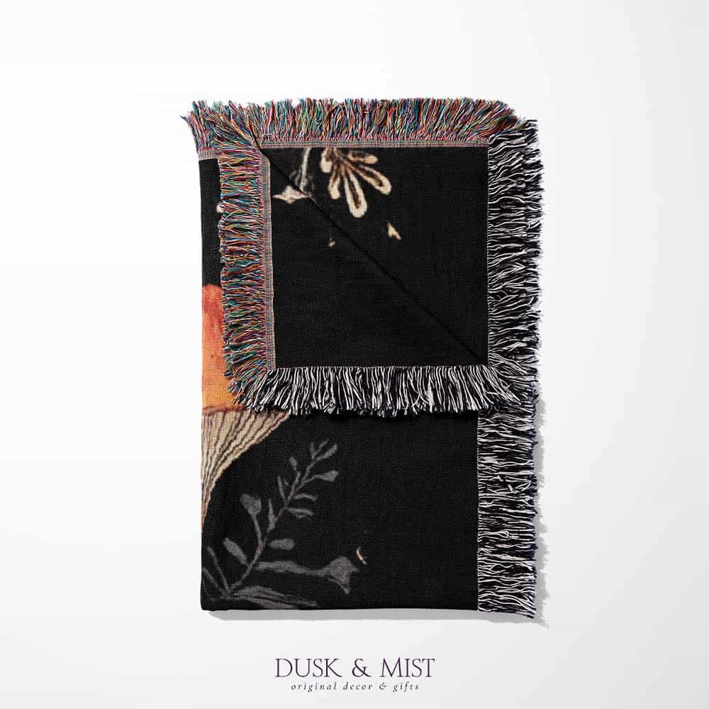 Woven cotton blanket showing a colorful cluster of mushrooms in red, gold, blue, and cream caps with surrounding plants and flowers on a black background.