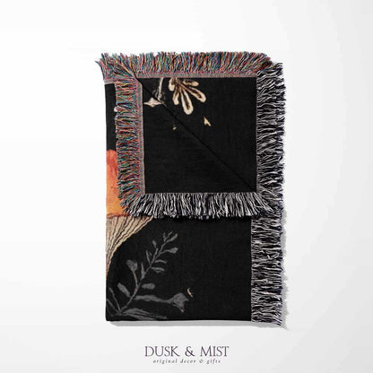 Woven cotton blanket showing a colorful cluster of mushrooms in red, gold, blue, and cream caps with surrounding plants and flowers on a black background.
