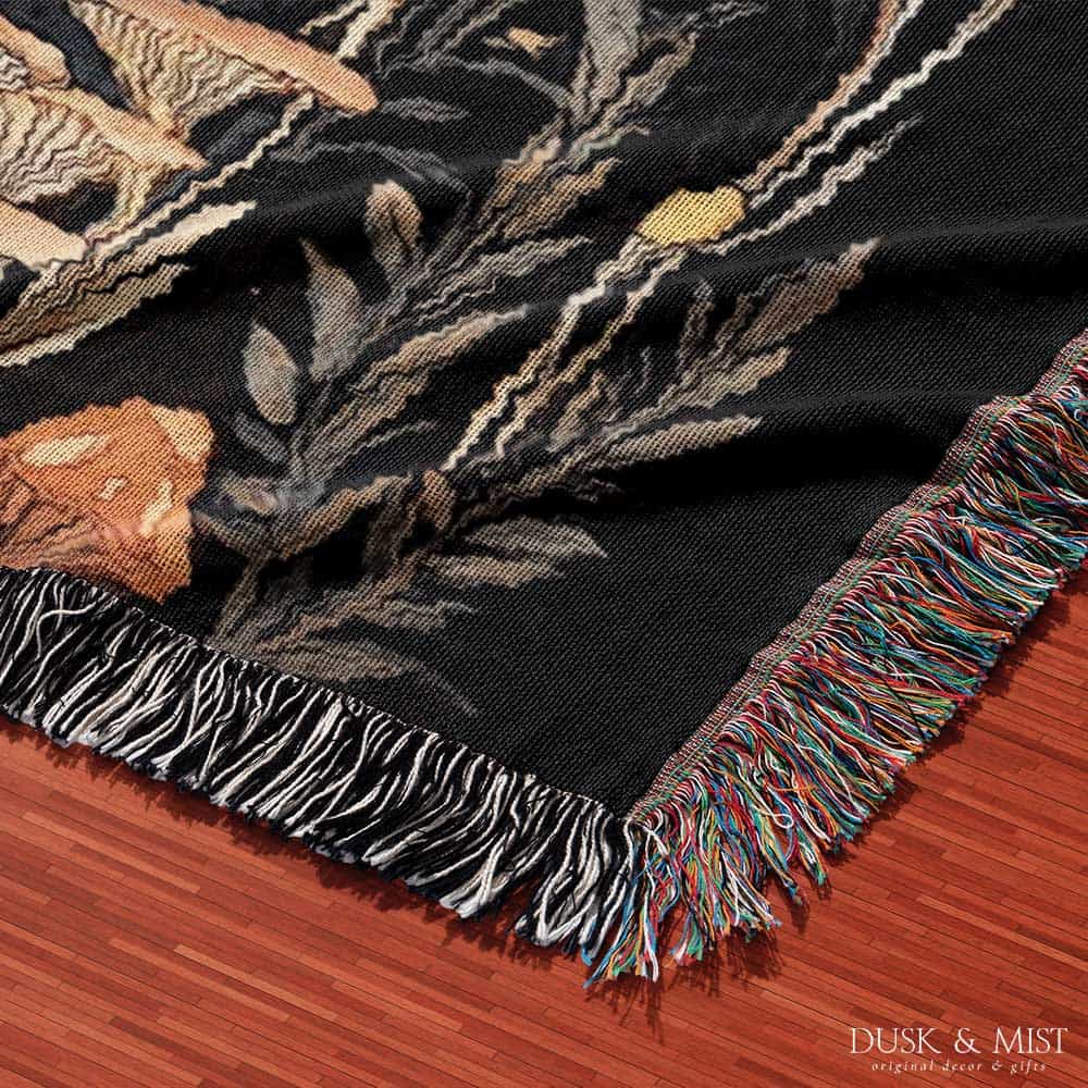 Woven cotton blanket showing a colorful cluster of mushrooms in red, gold, blue, and cream caps with surrounding plants and flowers on a black background.