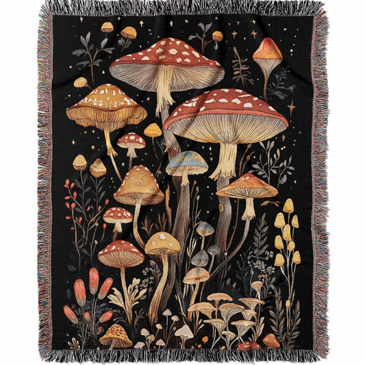 Woven cotton blanket showing a colorful cluster of mushrooms in red, gold, blue, and cream caps with surrounding plants and flowers on a black background.