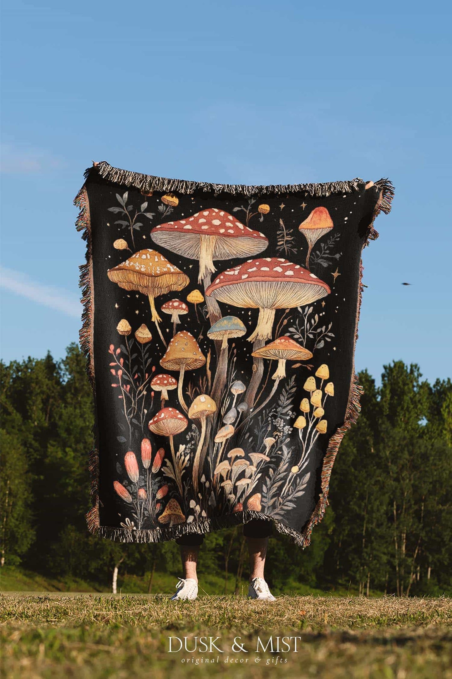Woven cotton blanket showing a colorful cluster of mushrooms in red, gold, blue, and cream caps with surrounding plants and flowers on a black background.