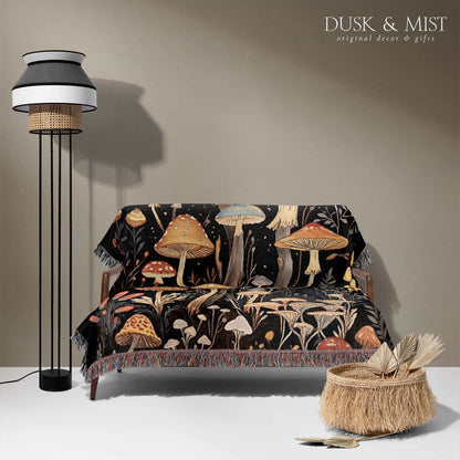 Woven cotton blanket showing a colorful cluster of mushrooms in red, gold, blue, and cream caps with surrounding plants and flowers on a black background.