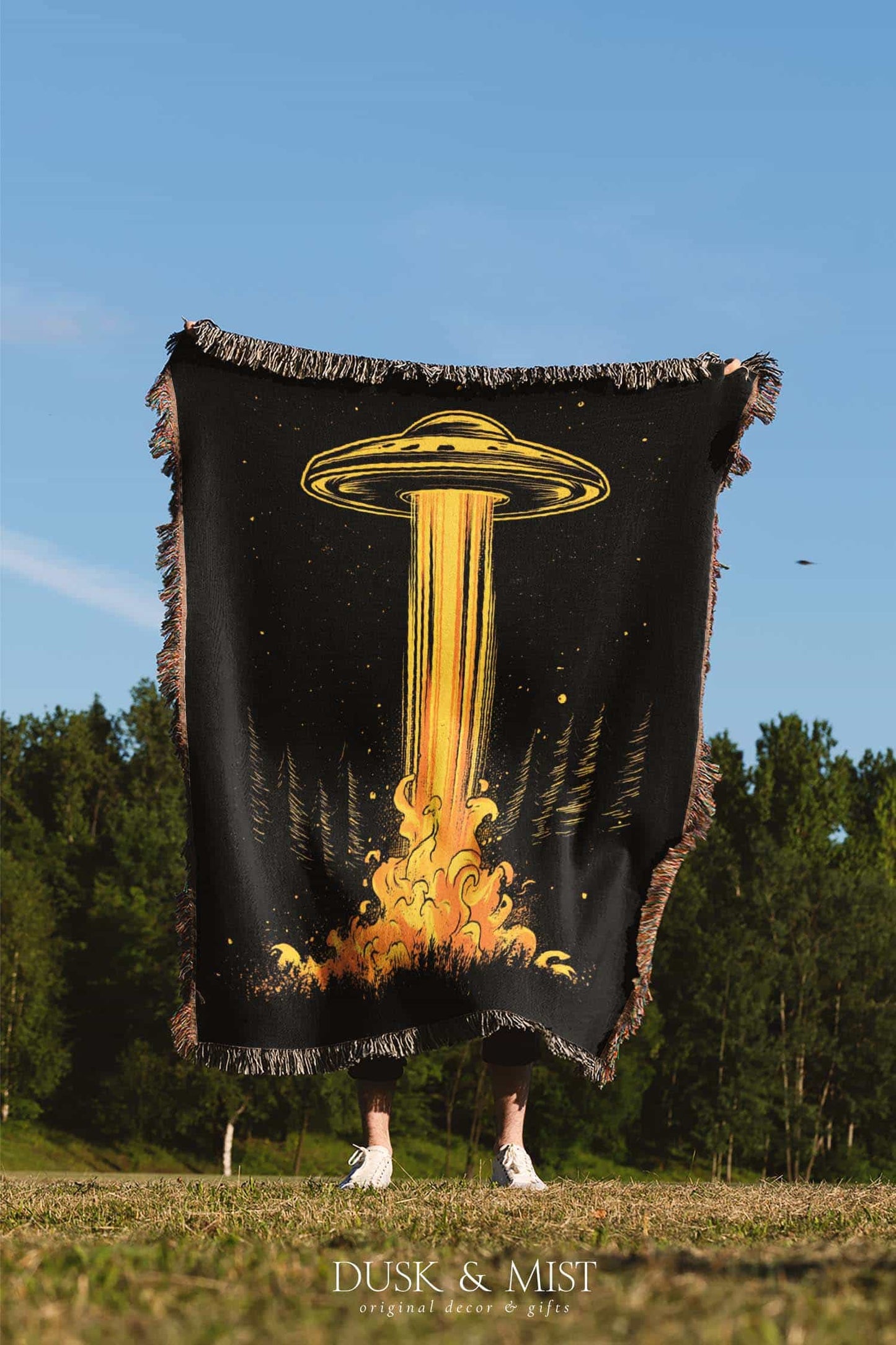 Woven cotton blanket featuring a UFO casting a golden beam into the forest at night, with glowing clouds and cosmic light effects.