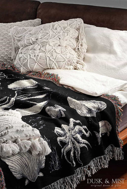 Woven cotton blanket featuring detailed ocean life illustration — whales, orca, turtles, jellyfish, octopus, seashells, fish, and coral on a black background.