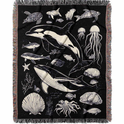 Woven cotton blanket featuring detailed ocean life illustration — whales, orca, turtles, jellyfish, octopus, seashells, fish, and coral on a black background.