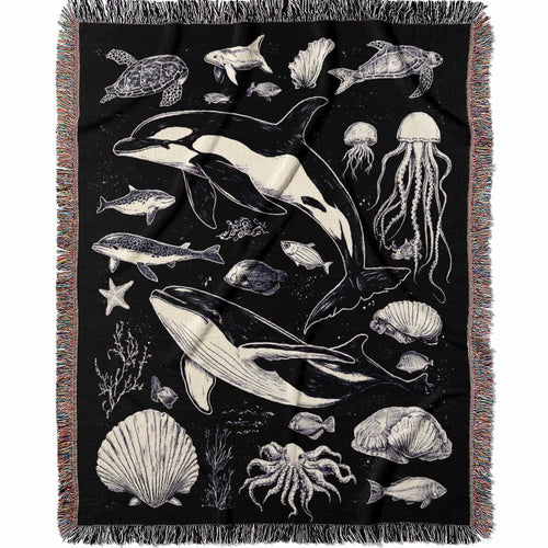 Woven cotton blanket featuring detailed ocean life illustration — whales, orca, turtles, jellyfish, octopus, seashells, fish, and coral on a black background.