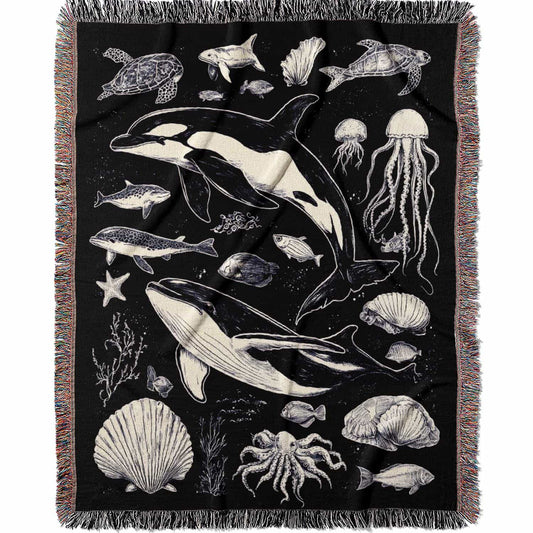 Woven cotton blanket featuring detailed ocean life illustration — whales, orca, turtles, jellyfish, octopus, seashells, fish, and coral on a black background.