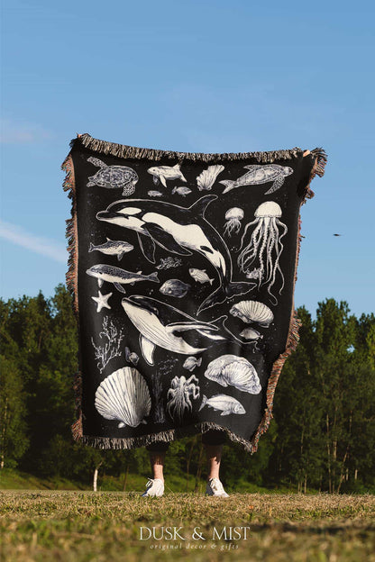 Woven cotton blanket featuring detailed ocean life illustration — whales, orca, turtles, jellyfish, octopus, seashells, fish, and coral on a black background.