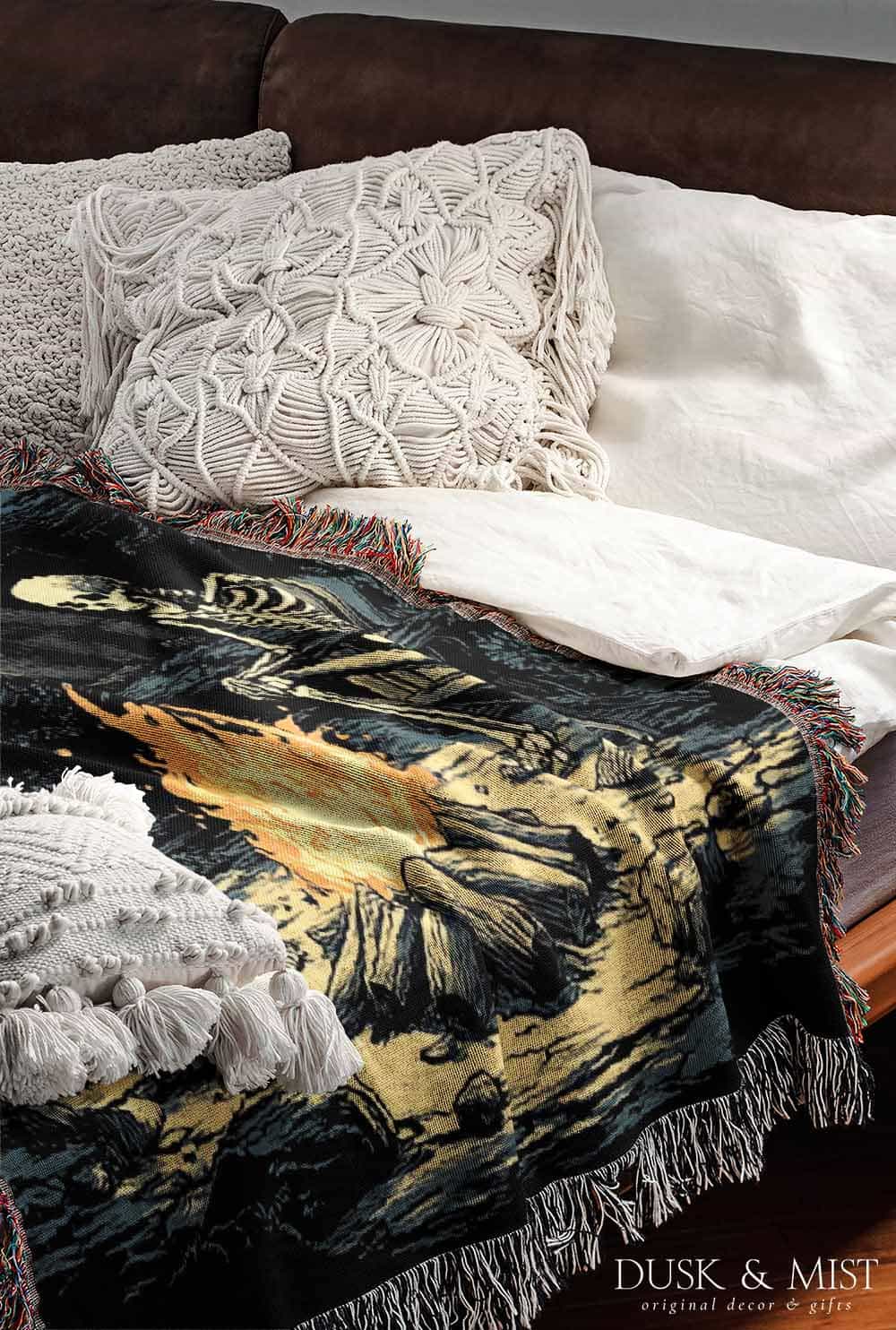Woven cotton blanket depicting two skeletons seated by a campfire in a forest clearing with a log cabin, full moon, and bear in the background.
