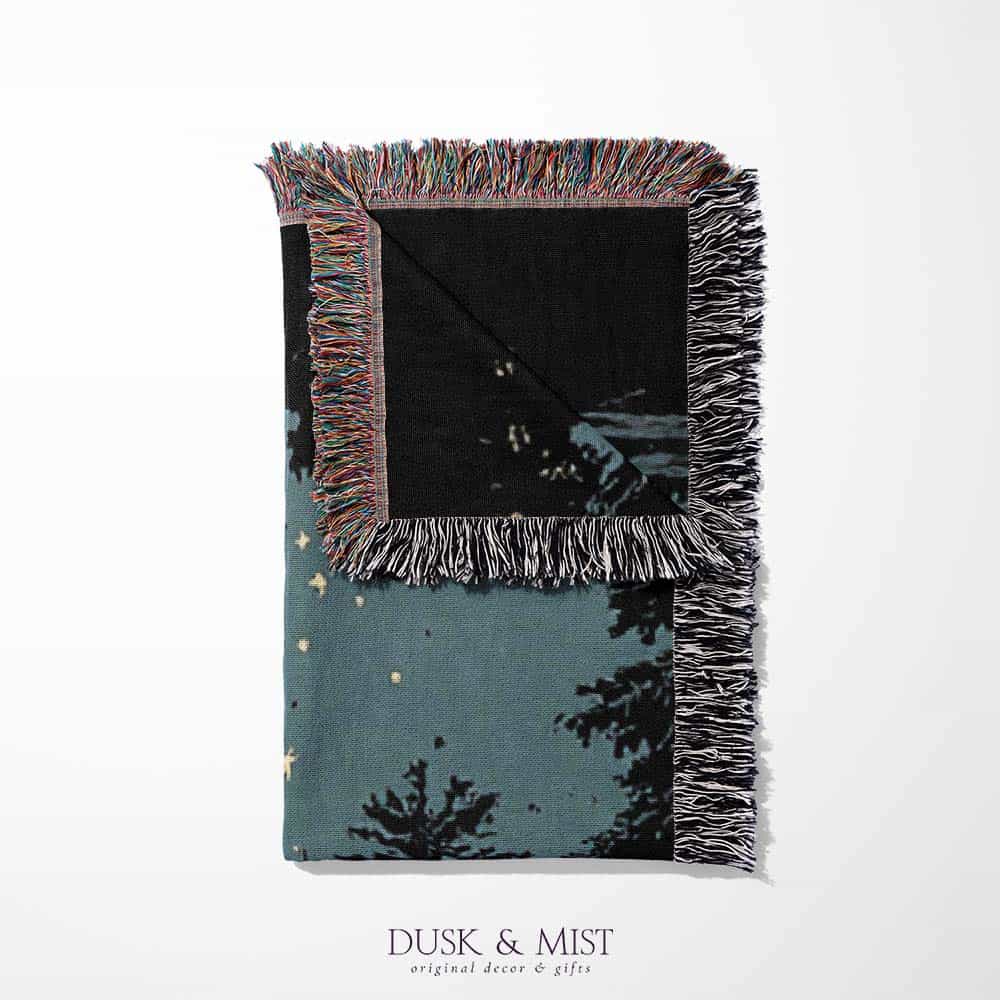 Woven cotton blanket depicting two skeletons seated by a campfire in a forest clearing with a log cabin, full moon, and bear in the background.