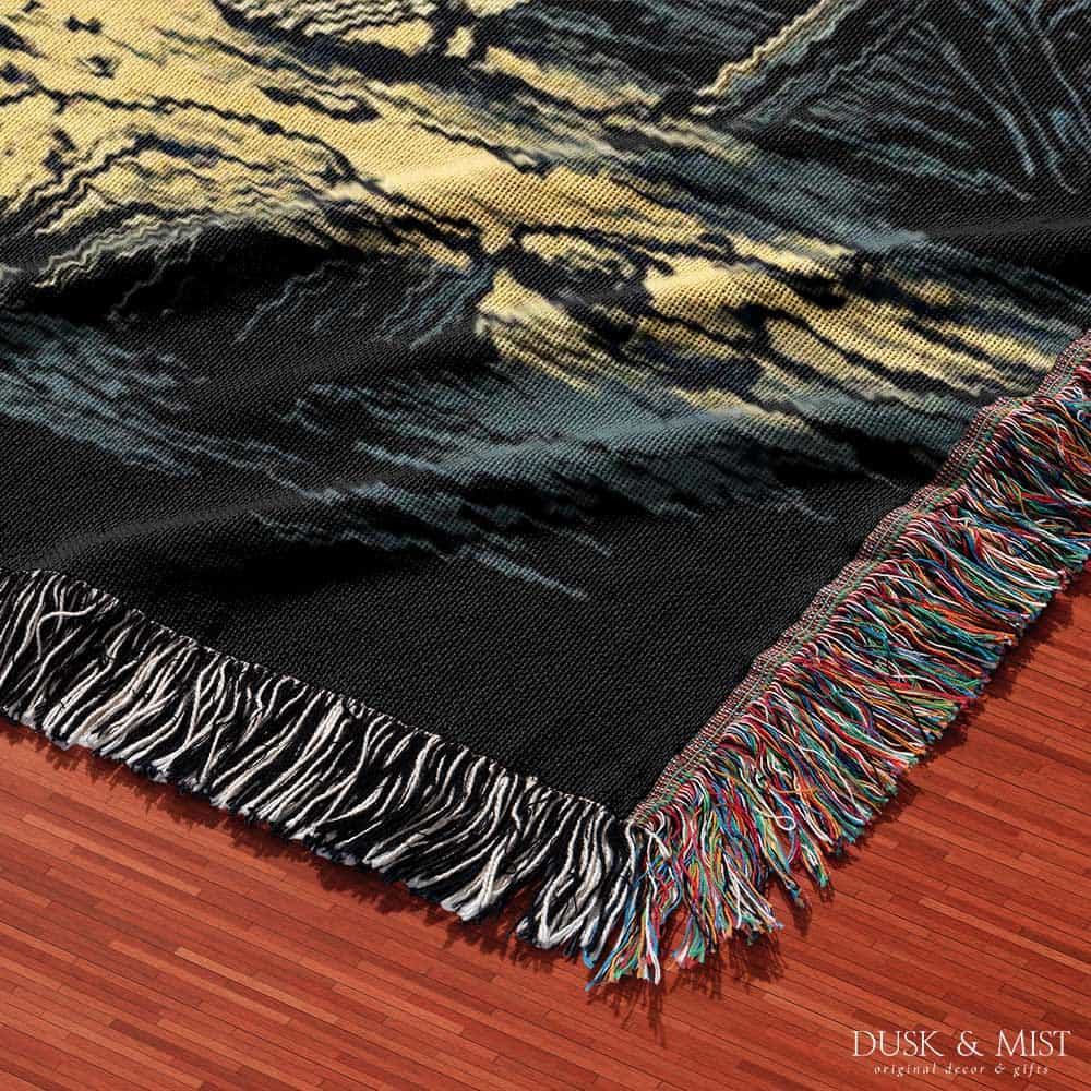 Woven cotton blanket depicting two skeletons seated by a campfire in a forest clearing with a log cabin, full moon, and bear in the background.