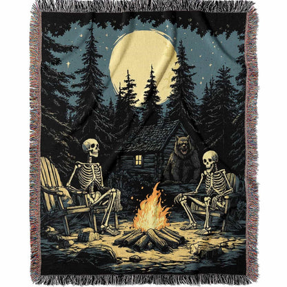 Woven cotton blanket depicting two skeletons seated by a campfire in a forest clearing with a log cabin, full moon, and bear in the background.