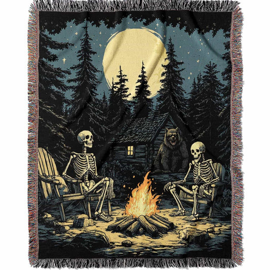 Woven cotton blanket depicting two skeletons seated by a campfire in a forest clearing with a log cabin, full moon, and bear in the background.