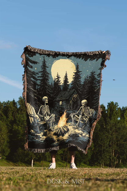 Woven cotton blanket depicting two skeletons seated by a campfire in a forest clearing with a log cabin, full moon, and bear in the background.
