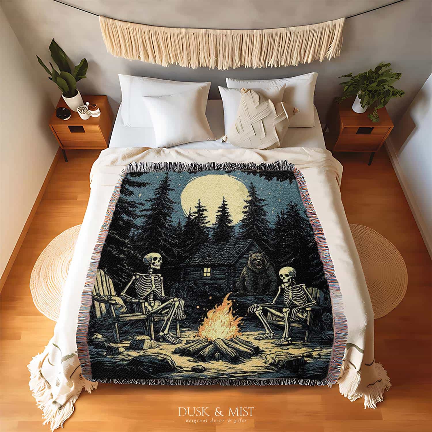 Woven cotton blanket depicting two skeletons seated by a campfire in a forest clearing with a log cabin, full moon, and bear in the background.