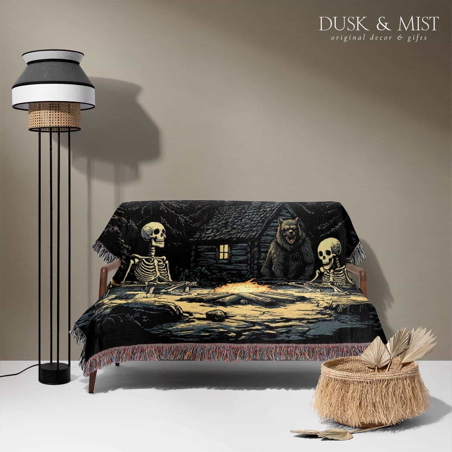 Woven cotton blanket depicting two skeletons seated by a campfire in a forest clearing with a log cabin, full moon, and bear in the background.