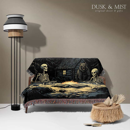Woven cotton blanket depicting two skeletons seated by a campfire in a forest clearing with a log cabin, full moon, and bear in the background.