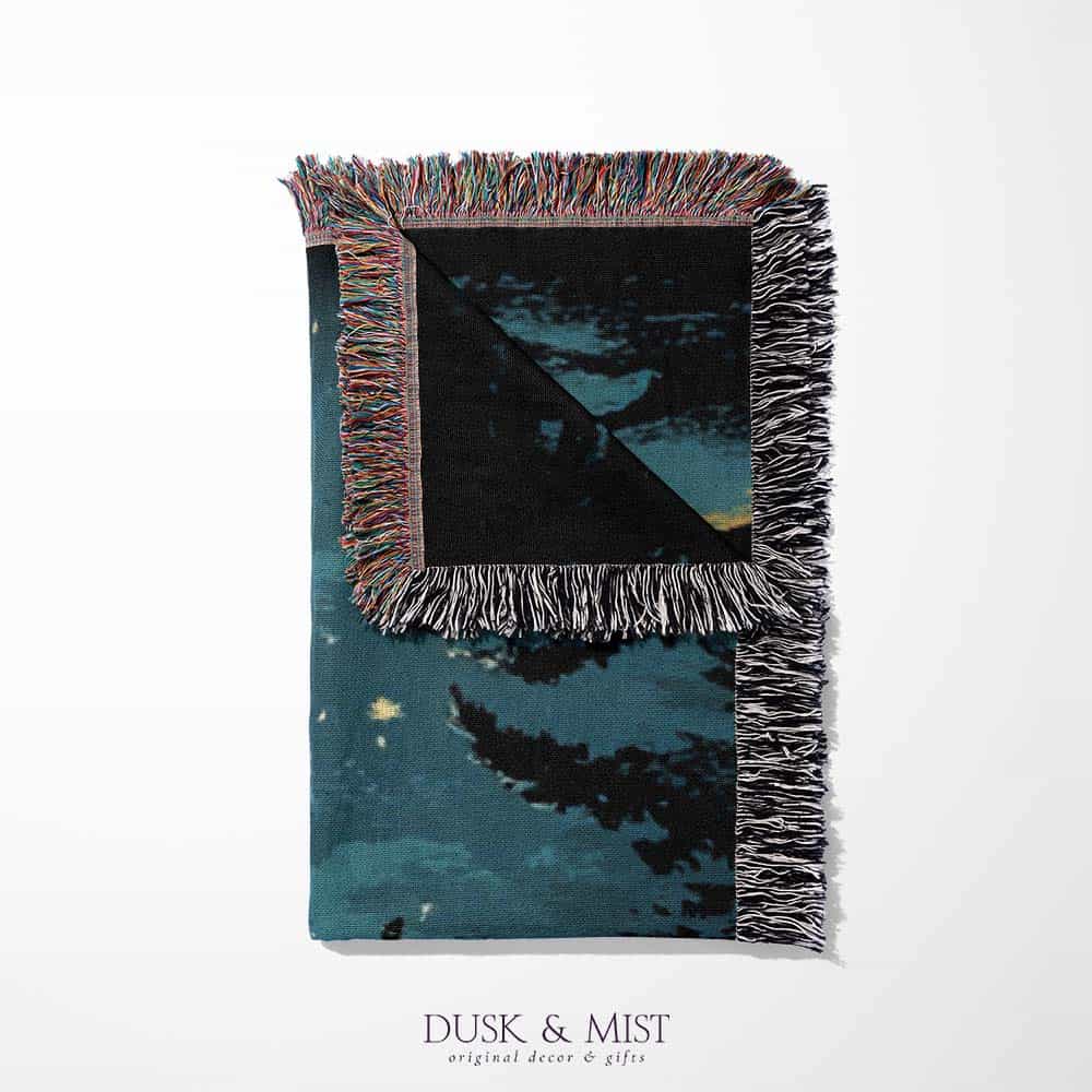 Woven cotton blanket showing two skeletons sitting around a campfire with a bear, log cabin, and full moon in a forest night scene.