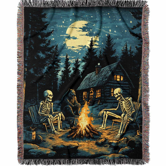 Woven cotton blanket showing two skeletons sitting around a campfire with a bear, log cabin, and full moon in a forest night scene.