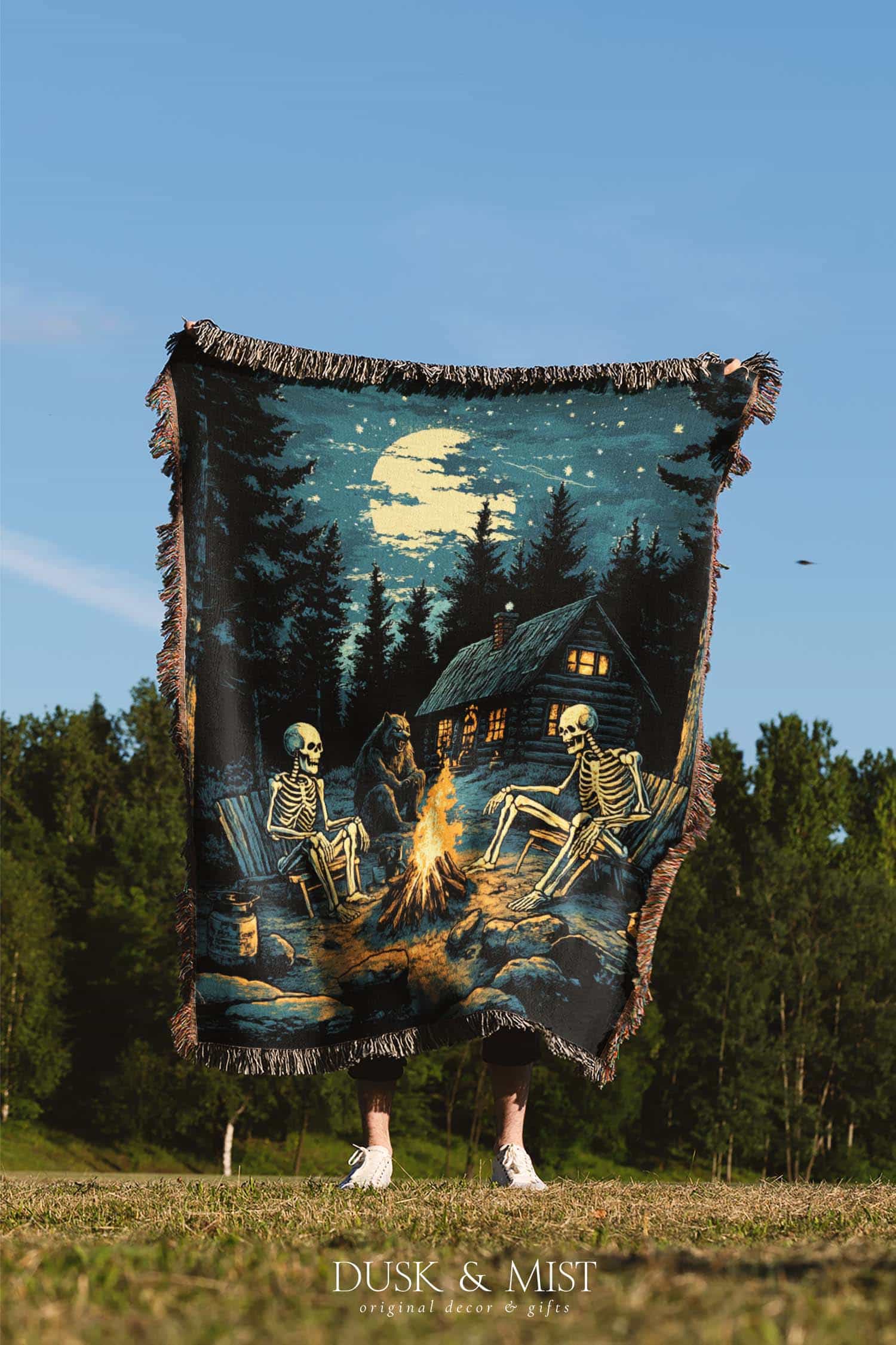 Woven cotton blanket showing two skeletons sitting around a campfire with a bear, log cabin, and full moon in a forest night scene.