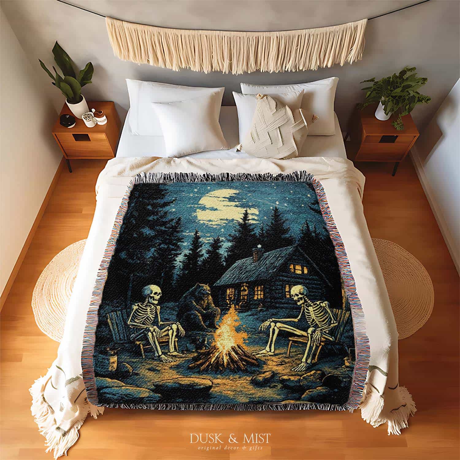 Woven cotton blanket showing two skeletons sitting around a campfire with a bear, log cabin, and full moon in a forest night scene.