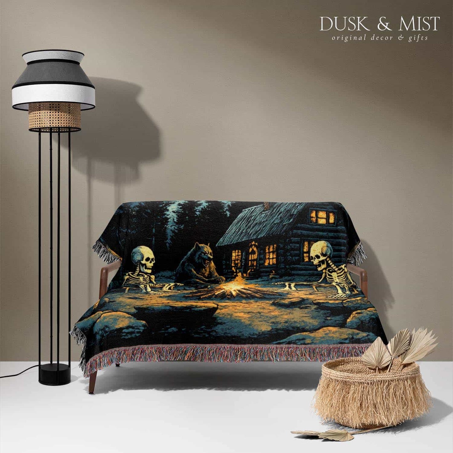 Woven cotton blanket showing two skeletons sitting around a campfire with a bear, log cabin, and full moon in a forest night scene.
