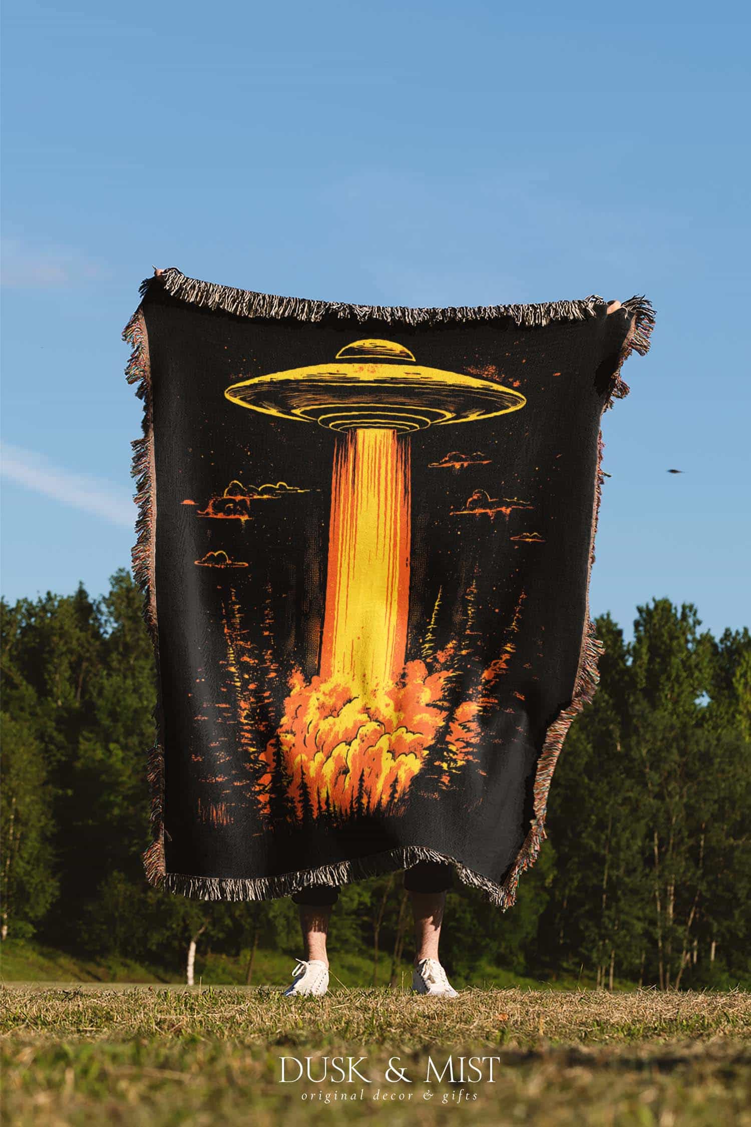 Woven cotton blanket featuring a UFO with a fiery orange beam illuminating a dark forest, with glowing clouds and night sky details.