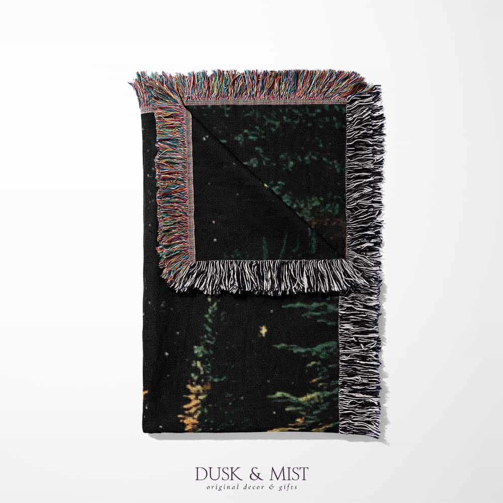 Woven cotton blanket showing a UFO shining a beam of light onto a cow in a forest clearing under a starry night sky.