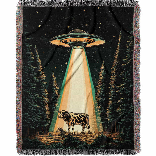 Woven cotton blanket showing a UFO shining a beam of light onto a cow in a forest clearing under a starry night sky.